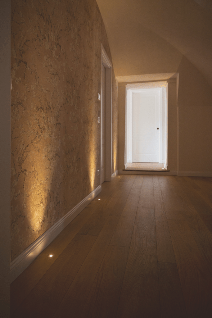 Aiace, pavimento in acacia bio in un ambiente dallo stile classico con carta da parati floreale, illuminato da faretti incassati nel parquet.